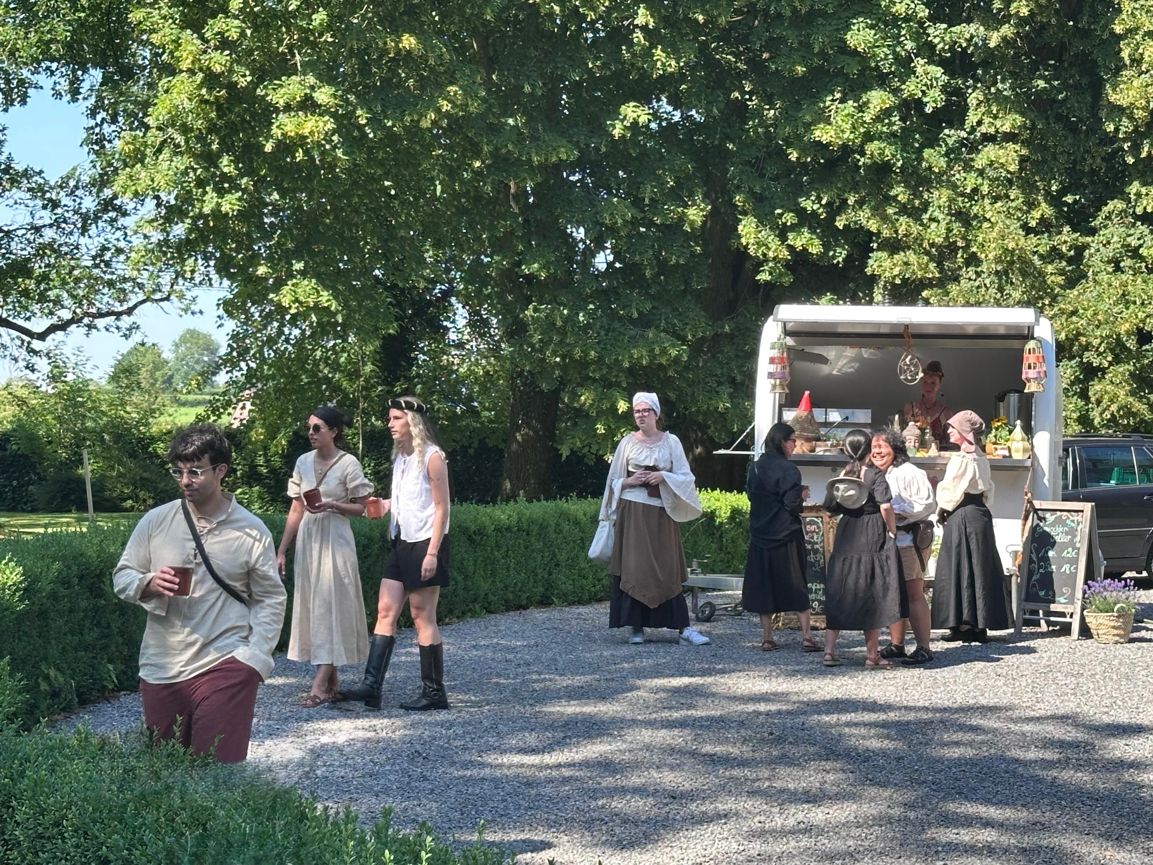 Marktstand und Gäste in Gewändern auf dem Kiesweg